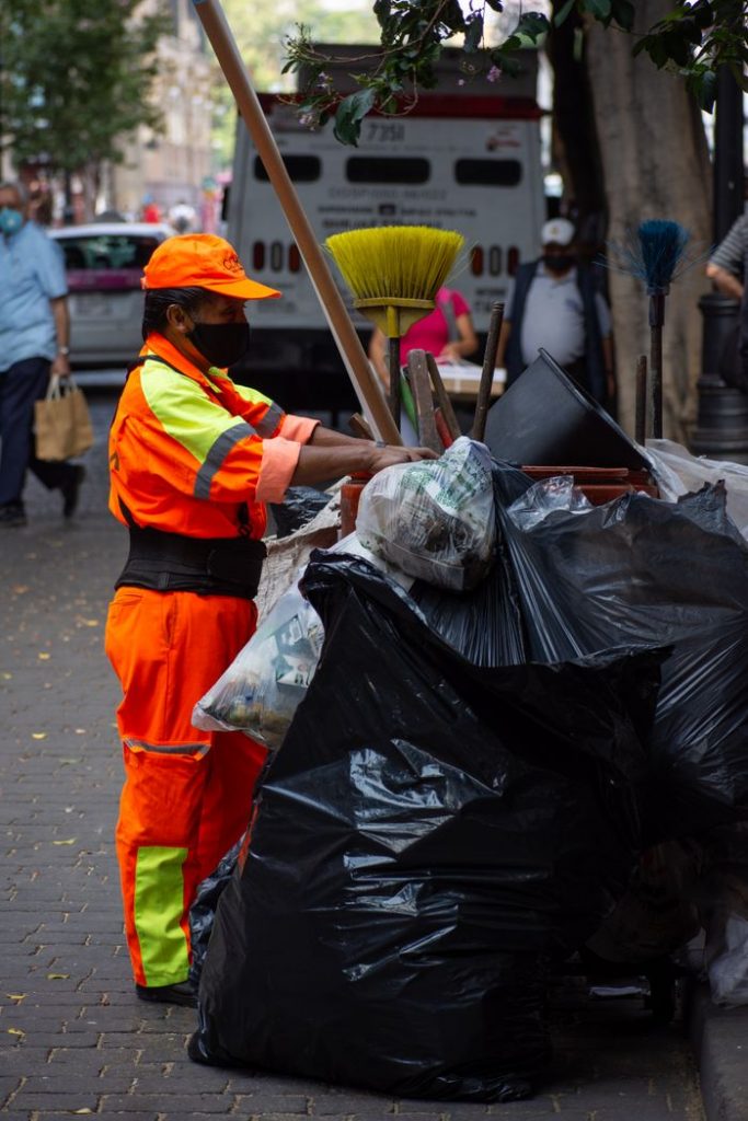 A city worker in orange uniform cleaning streets, managing trash on an urban sidewalk.