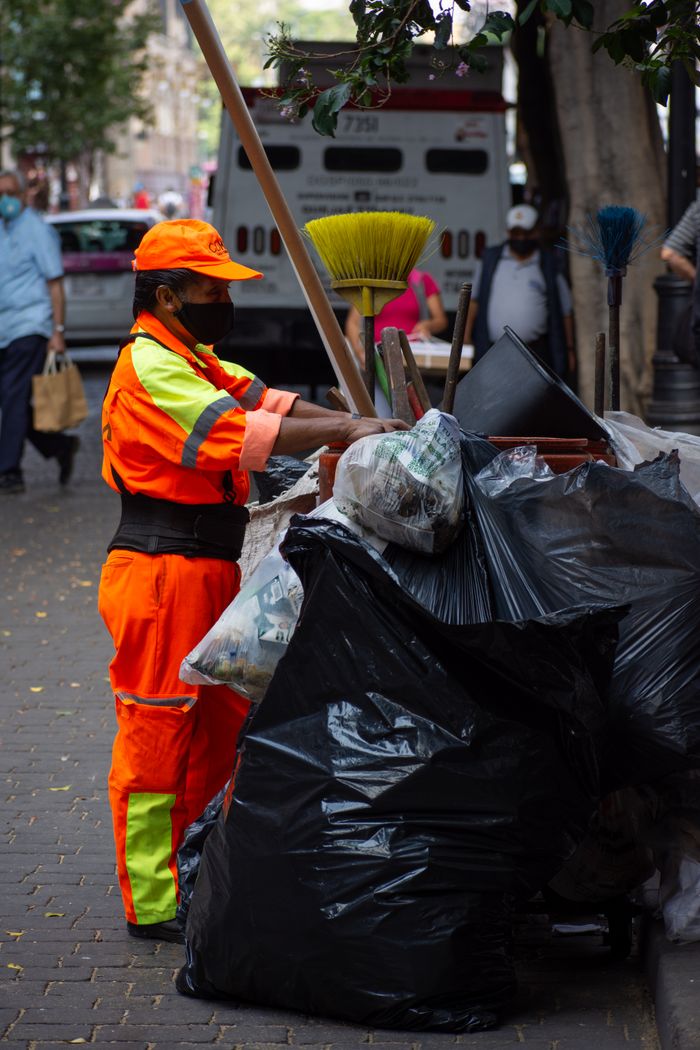 A city worker in orange uniform cleaning streets, managing trash on an urban sidewalk.